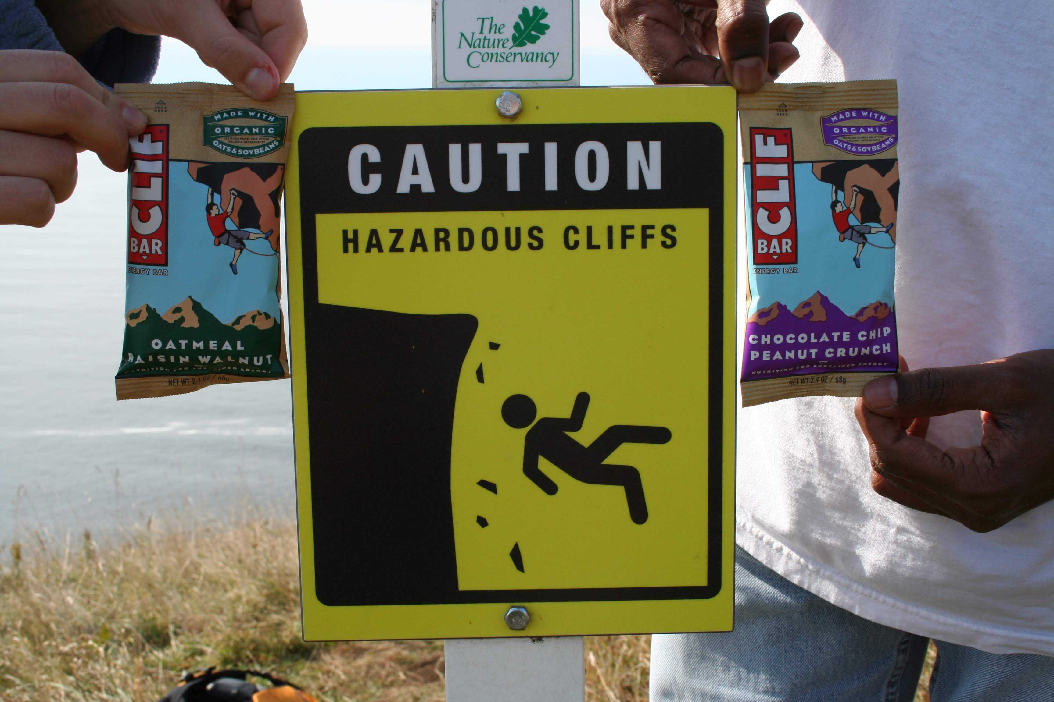 A sign that reads "Hazardous Cliffs" with Sam and Elyssia holding up
       Cliff Bars on either side of it